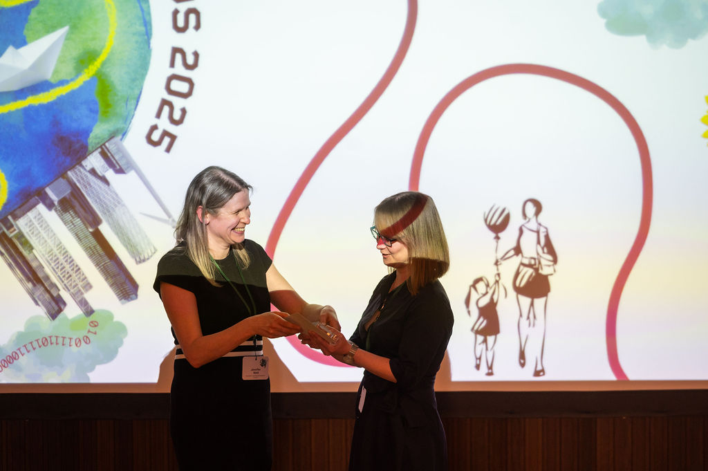 Dr Jen Gold smiles as she hands over the Emerging Impact Award trophy to Dr Jennifer Seddon in front of a screen with award branding