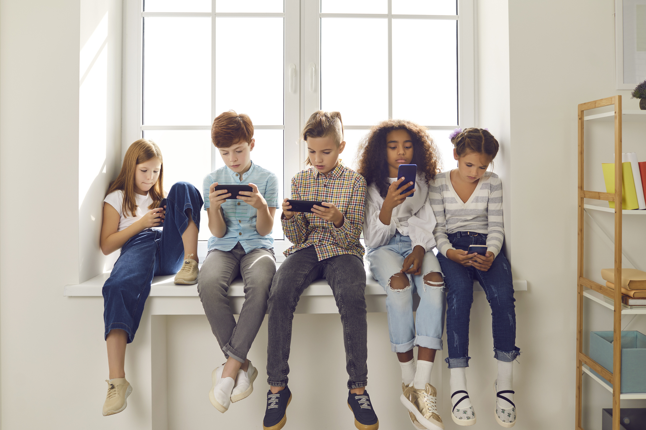 Group of children of different nationalities sit in a row on the windowsill and with lack of emotion play online games or read social networks on mobile phones