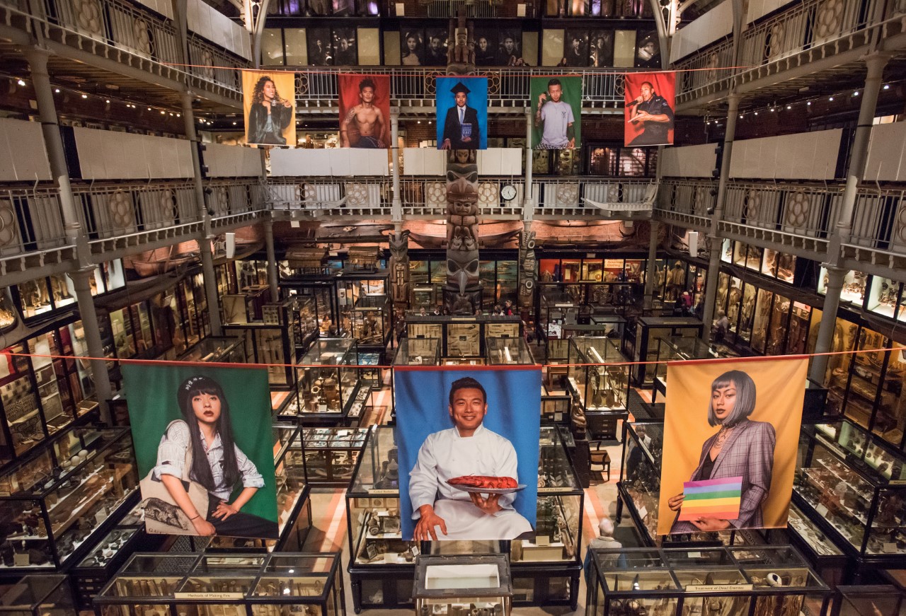 Panoramic view of the Performing Tibetan Identities exhibition inside the Pitt Rivers Museum. Large, photographic banners displaying people from the Tibetan community in in different styles of dress hang over the museum displays.