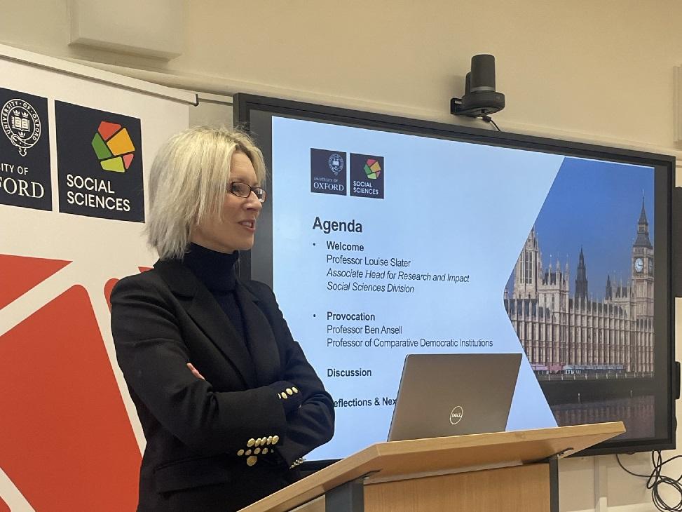 Professor Louise Slate stands at a lecture podium in front of Social Sciences Divisional branding and a slide of the agenda for the Democracy seminar