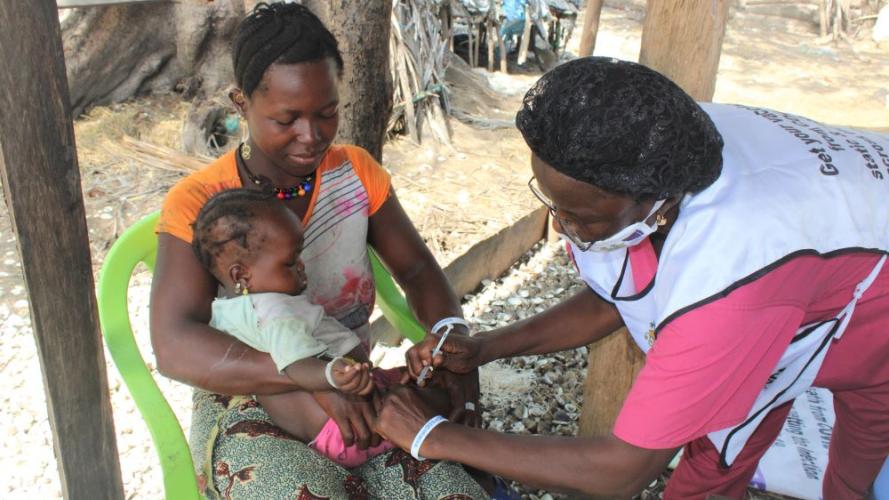 A young Sierra Leonean woman sits in an open-air clinic with a baby on her lap, whilst a female nurse administers an injection into the baby's leg. 