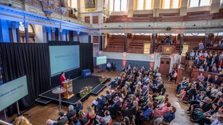 An audience in the Sheldonian Theatre. They are facing a stage, where the Vice-Chancellor is speaking at a podium, wearing a red suit. 