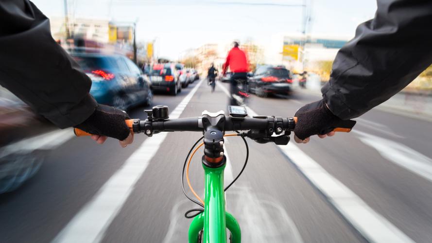 View from the handlebars of a cyclist in the middle of a busy road. Cars and others cyclists around them are blurred.