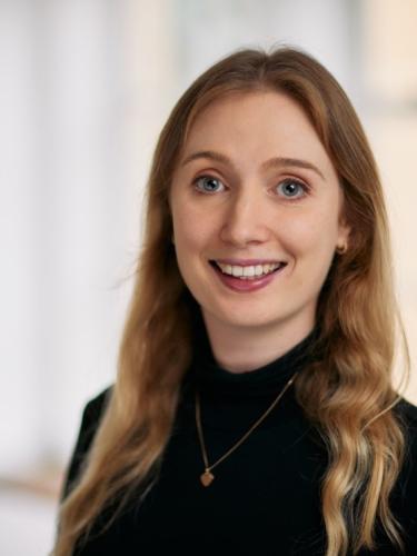 Dr. Chloe Bracegirdle is smiling in front of a white background. She has dark blonde hair and is wearing a black polo neck sweater.