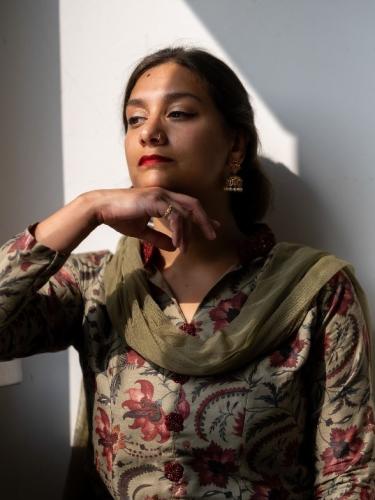 Dr. Shyama Vermeersch is wearing striking red lipstick and is looking off to the side. She is also wearing a green scarf and has dark brown hair tied back.