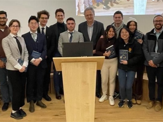 Seventeen smiling, smartly dressed people stand behind a lectern, some holding copies of a book. 