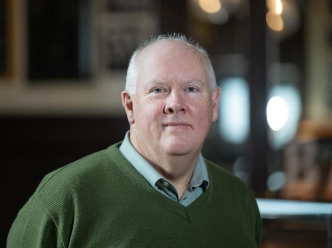 Professor Timothy Power has short white hair and is wearing a green jumper and light blue shirt