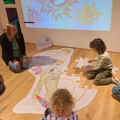 Two children sit on the floor to draw on a poster with an adult