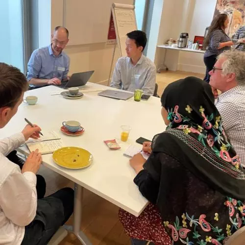 Researchers seated around a table are in conversation; one is typing, two others are taking notes by hand. In the background is a flipchart and more people in conversation