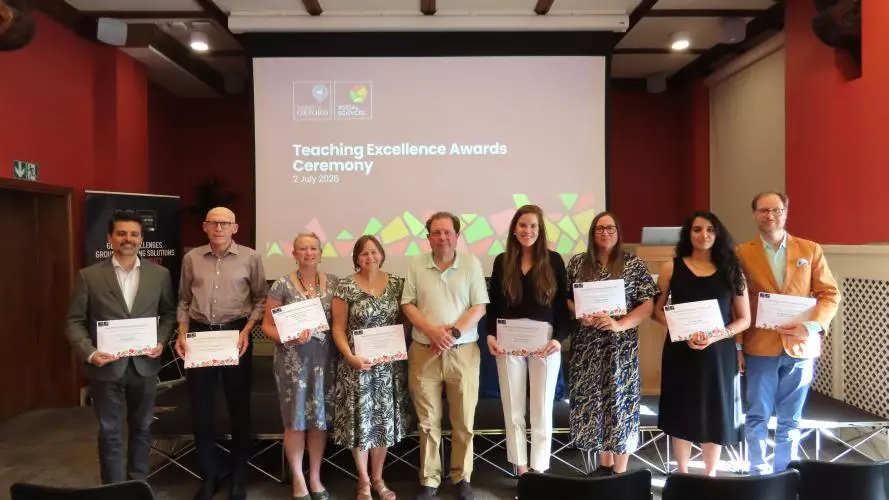 A group of 9 academics, standing in a line and holding certificates in front of a projected slide that says 'Teaching Excellence Awards ceremony'