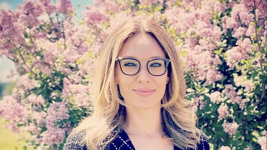 Celeste has long blonde hair, is wearing glasses and smiling into the camera
