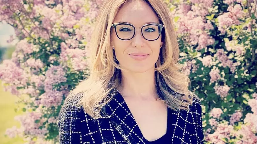 Celeste has long blonde hair, is wearing glasses and smiling into the camera