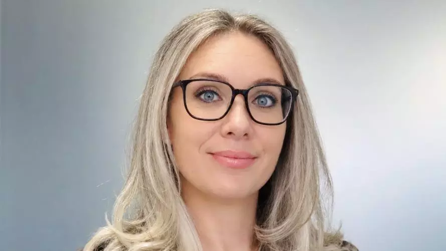 Celeste has long blonde hair, is wearing glasses and smiling into the camera