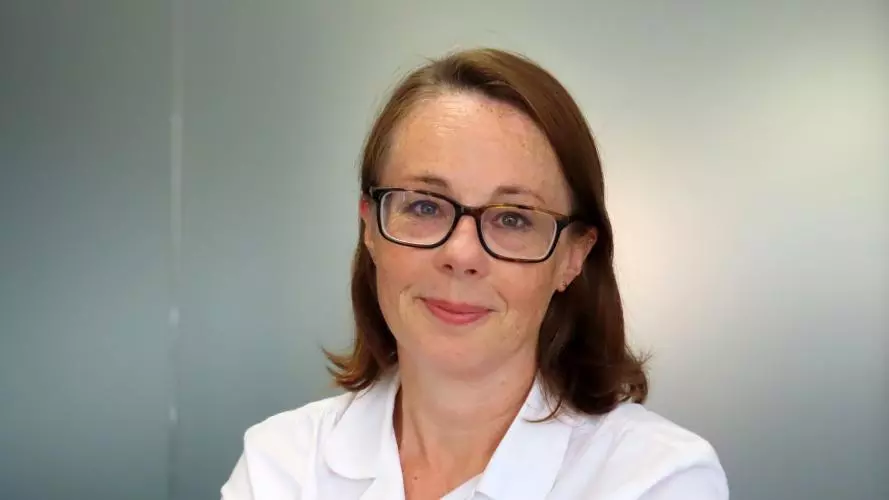 Rachel has shoulder-length brown hair and dark glasses. She is wearing a white shirt.