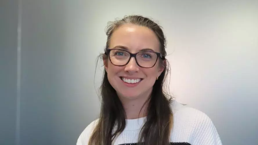Sophie has long brown hair. She is wearing large round glasses and a white jumper with thin black stripes