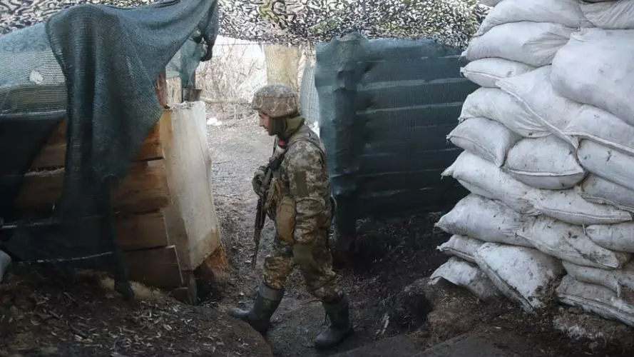 A Ukrainian soldier dressed in camouflage uniform and holding a gun pointed towards the ground patrols a post fortified with sandbags and draped in camouflage.