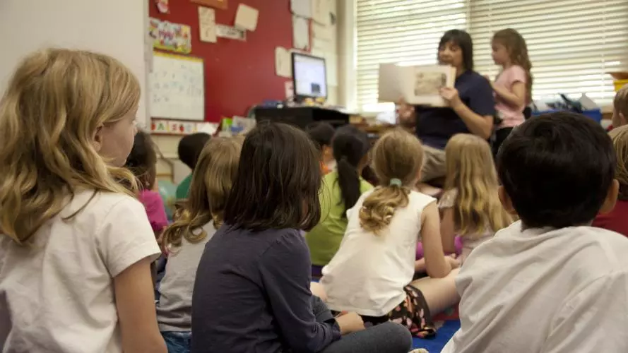 Taken from the back of a classroom, where a primary school teacher reads from a book to a room full of young children, with their backs to the camera. All the children are watching as she holds the book facing outwards, so they can all see the pages