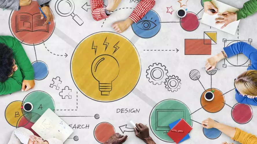 Eight people of diverse ethnicities sit around a table sketching out colour ideas in graphic form, including a book, lightbulb, eye, arrow, jigsaw puzzle piece, and cog.