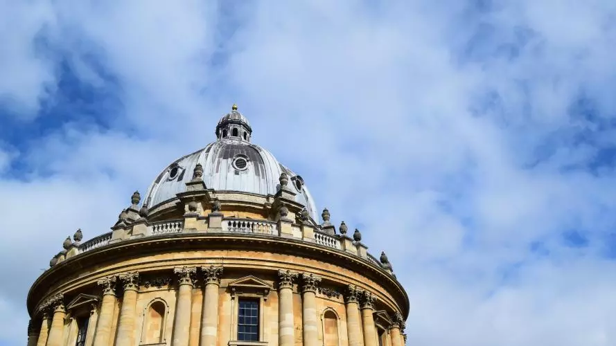 A view of the circular Radcliffe Camera library showing the blue-grey leaded roof and yellow standstone walls against a bright blue sky with whispy clouds