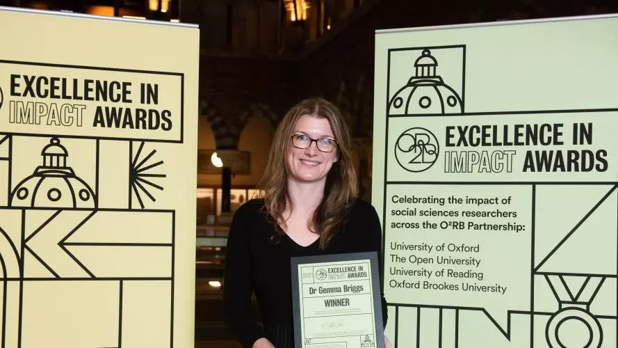 Gemma Briggs smiles as she holds her Impact Awards winners certificate in front of the O2RB Excellence in Impact Awards event branding