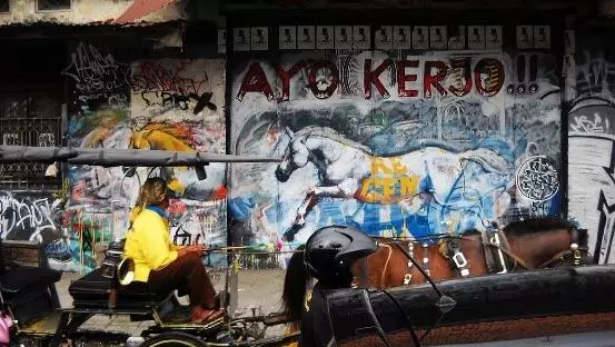 teenager in yellow t-shirt drives a horse drawn carriage down a street in indonesia