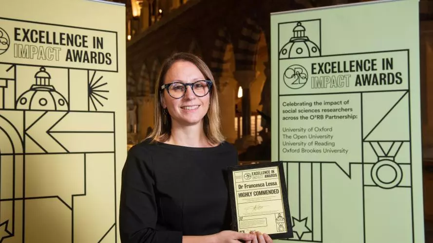 Dr Francesca Lessa holds her Excellence in Impact Awards Highly Commended certificate at the Awards reception evening, flanked by pull-up banners of the awards branding. In the background are the arches of the Museum of Natural History. The banners read: 