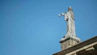A statue of a robed figure blowing a trumpet, positioned on the roof of a building against a bright blue sky