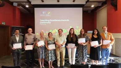 A group of 9 academics, standing in a line and holding certificates in front of a projected slide that says 'Teaching Excellence Awards ceremony'