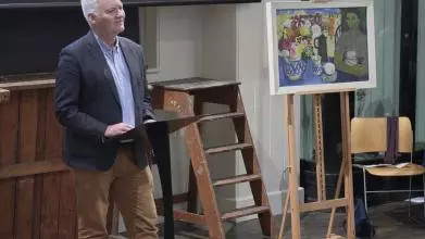 Associate Professor Ian Thompson is standing in front of a ladder and some art. He is wearing brown trousers, blue shirt and navy blazer and presenting to the room. 