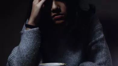 A young woman sits in a dark room with her head in one hand, leaning over a cup of tea