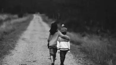 A black and white photo of two small children walking down a track, away from the camera. The bigger child has their arm around the smaller one, as if comforting them.