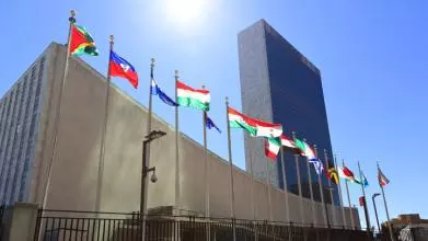 The UN headquarters against a bright blue sky, with a row of national flags in the foreground