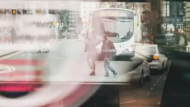 Photo of a bus window reflecting people walking and a bus and cars driving through a street. The image is slightly blurred and gives the impression of movement.