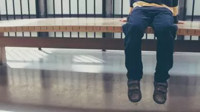 Shows a young child sitting alone on a bench, their feet dangling above the floor. The child's face is not visible.