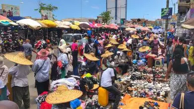 A busy market scene where people are buying and holding clothes and shoes. Many people are wearing sunhats and the market stalls are shaded by sun umbrellas.