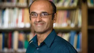 Portrait photo of Professor Yadvinder Malhi, smiling against a blurred background of shelves full of books