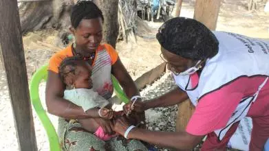A young Sierra Leonean woman sits in an open-air clinic with a baby on her lap, whilst a female nurse administers an injection into the baby's leg. 
