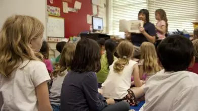 Taken from the back of a classroom, where a primary school teacher reads from a book to a room full of young children, with their backs to the camera. All the children are watching as she holds the book facing outwards, so they can all see the pages
