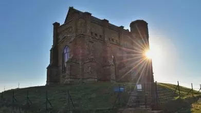 Looking uphill towards English Heritage's 14th-century St Catherine's Chapel on a hilltop in Abbotsbury, Dorset. The sky behind is blue and the dazzling sun is just peering around the edge of the chapel. The chapel is surrounded by a protective fence.