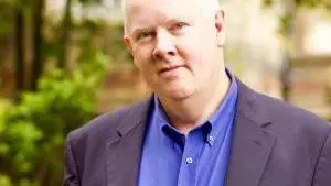 A white man with grey hair stands in a garden - the background is blurred. He wears a bright blue shirt and dark blue jacket.