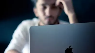 A young man sits in front of his computer screen, his hand held up to his head