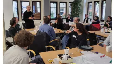 A male academic addresses a room full of researchers who listening to him attentively, and seated around tables that are covered in pens and paper, brainstorming activities, and lunch