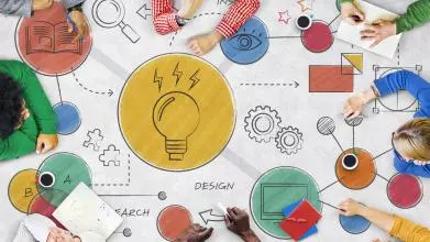 Eight people of diverse ethnicities sit around a table sketching out colour ideas in graphic form, including a book, lightbulb, eye, arrow, jigsaw puzzle piece, and cog.