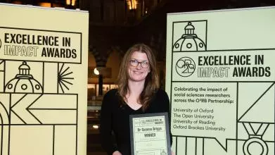 Gemma Briggs smiles as she holds her Impact Awards winners certificate in front of the O2RB Excellence in Impact Awards event branding