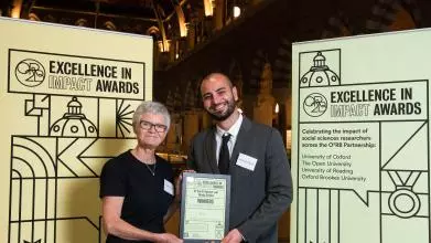  Dr Sarah Spencer and Mr Nicola Delvino smile as they hold their Impact Awards winners certificate, in front of the O2RB Excellence in Impact Awards event branding