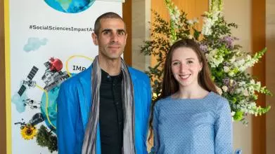 Dr Six Silberman and Aislinn Kelly-Lyth smile to camera.  In the background is the Impact Awards branding and a flower arrangement.