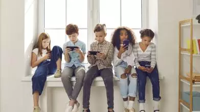 Group of children of different nationalities sit in a row on the windowsill and with lack of emotion play online games or read social networks on mobile phones