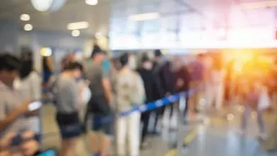 Blurred image of a queue of people at an airport's border control. The is going down through a window in the background.