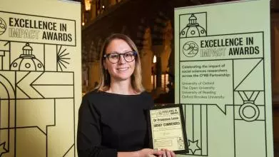 Dr Francesca Lessa holds her Excellence in Impact Awards Highly Commended certificate at the Awards reception evening, flanked by pull-up banners of the awards branding. In the background are the arches of the Museum of Natural History. The banners read: 