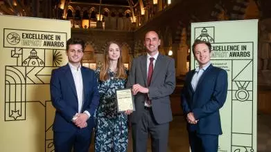 Mr Dan Snow, Ms Martha Kirby, Dr Stephen Fisher and Dr Zack Grant smile as they hold their Highly Commended certificate, flanked by impact awards branding at the Museum of Natural History